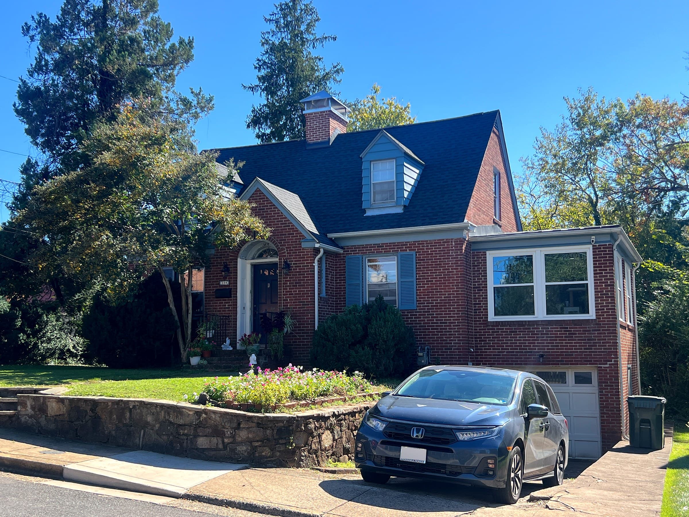 Brick Cape Cod-style home in Culpeper, VA with a blue-gray dormer, blue shutters, an arched front entry, a stone garden wall with flowers, a minivan in the driveway, and a GAF Timberline HDZ Charcoal roof