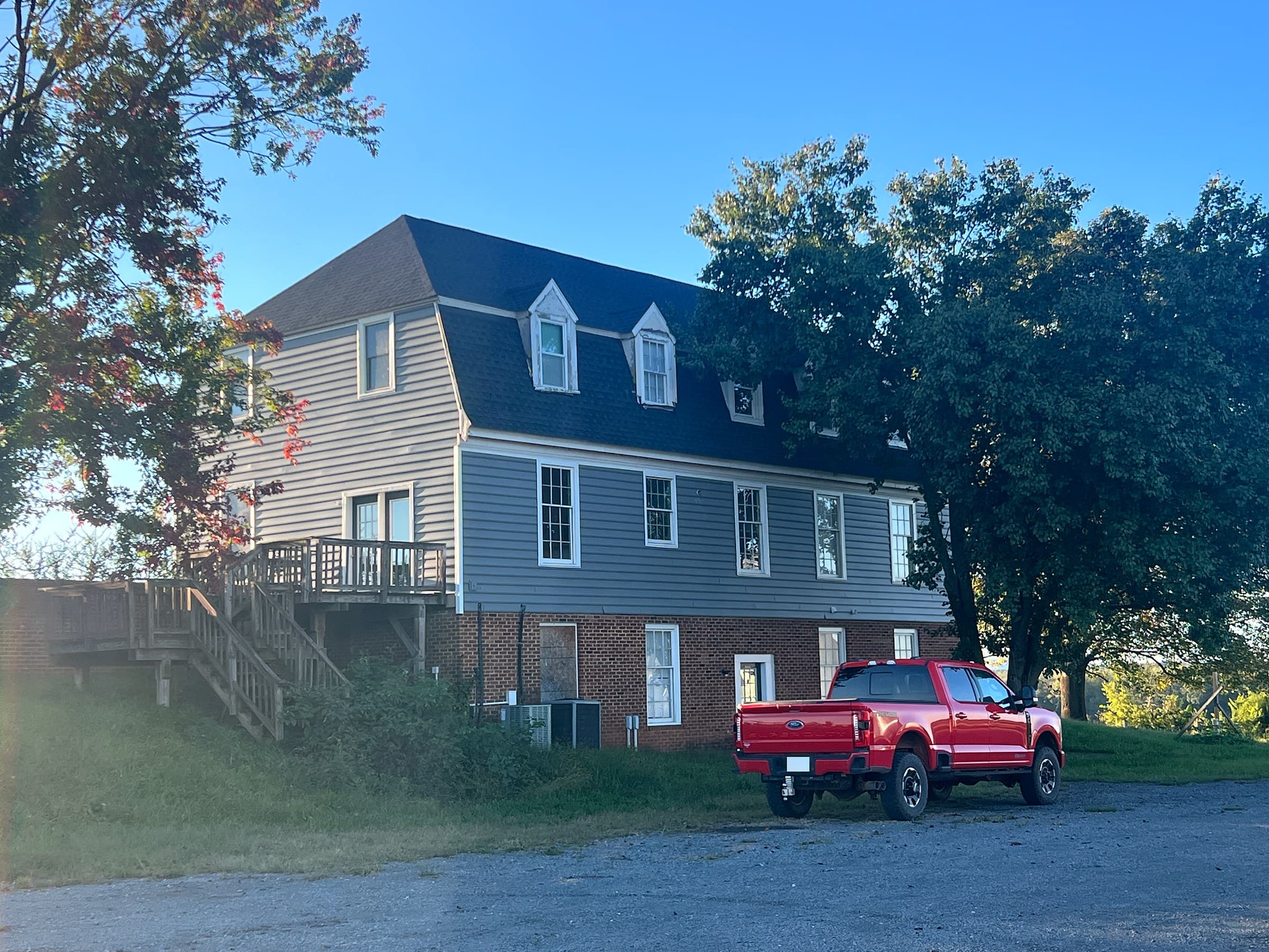Large gambrel-roof farmhouse in Culpeper, VA with dormers, gray siding over a brick lower level, a wooden deck with stairs, a shade tree, a red pickup in the gravel lot, and a GAF Timberline HDZ Charcoal roof